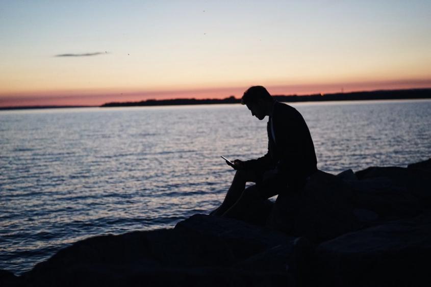 Hommeassis avec téléphone premier plan assis en contre-jour fond coucher soleil bord de mer