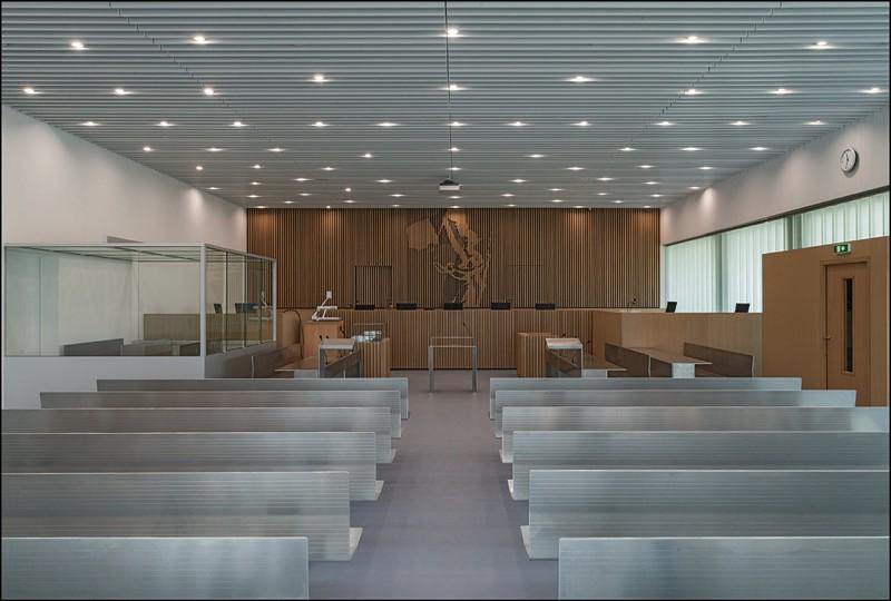 Salle d’audience dans laquelle les espaces sont clairement identifiés par l’estrade en bois du jury, le box des détenus, le pupitre et les assises des parties en présence, et les bancs en aluminium pour le public.