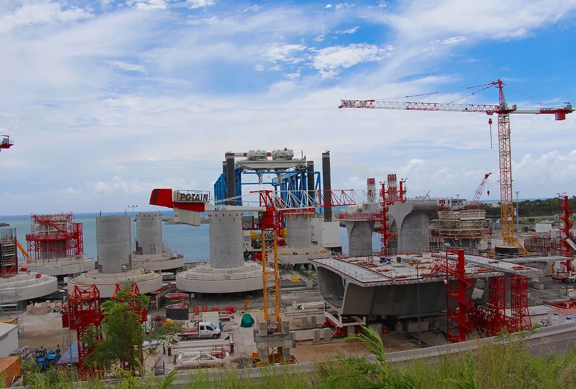 Une fois leur phase de maturation terminée, les embases et têtes de piles sont embarquées directement depuis l’aire de préfabrication sur la barge (au fond en bleu) qui les mettra en place. Une fois leur phase de maturation terminée, les embases et têtes de piles sont embarquées directement depuis l’aire de préfabrication sur la barge (au fond en bleu) qui les mettra en place.