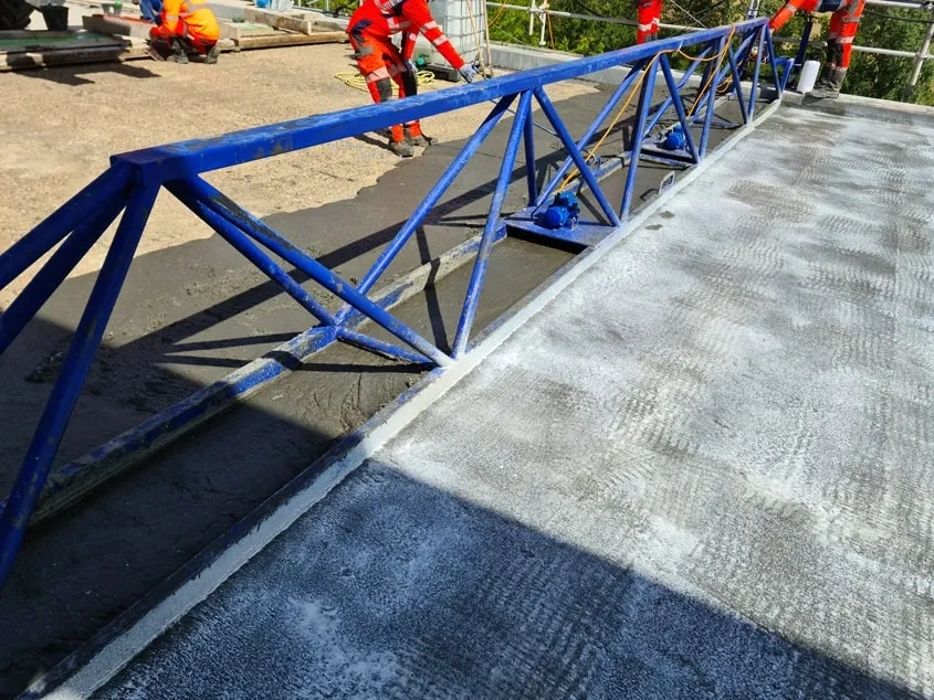 Coulage de la couche d’étanchéité BFUP du tablier (photo Ingerop). Une fois coulé sur le support béton humidifié, le BFUP est légèrement vibré à l’aide d’une règle vibrante. Cette opération permet d’étaler le matériau et de fixer son épaisseur selon une pente transversale de 2 %. Une fois mis en œuvre, le BFUP est immédiatement traité avec un produit de cure puis recouvert d’un géotextile. Celui-ci est maintenu humide pendant quatre jours, afin de garantir une prise homogène du BFUP.