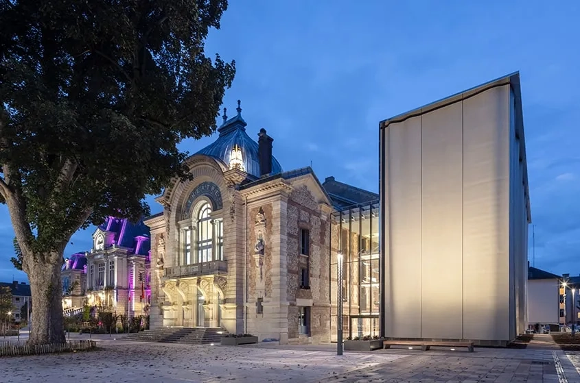 Dans l’éclairage nocturne, le dialogue entre le patrimoine et l’architecture contemporaine gagne une forme d’emphase qui théâtralise le lieu depuis l’espace public et met le spectateur en condition.