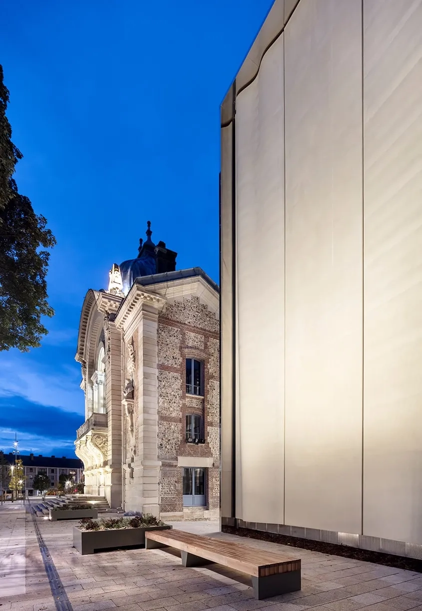 Dans l’éclairage nocturne, le dialogue entre le patrimoine et l’architecture contemporaine gagne une forme d’emphase qui théâtralise le lieu depuis l’espace public et met le spectateur en condition.