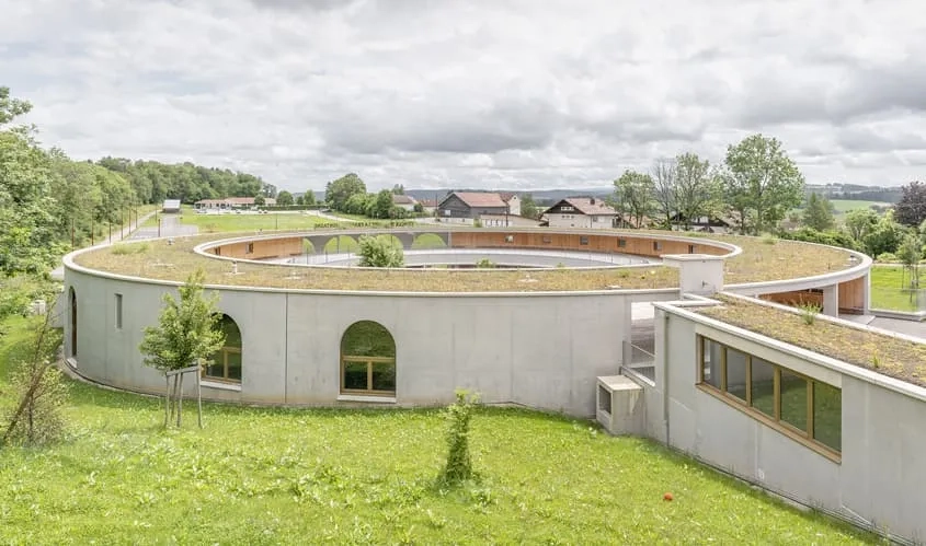 Encastrée dans le talus, l’extension ceinte par des murs de béton brut, sobres. La toiture végétalisée contribue à son insertion paysagère.