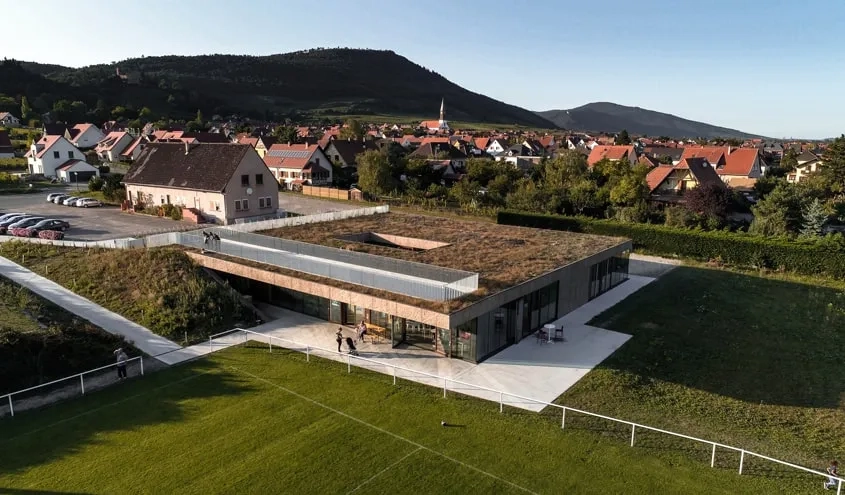 La maison de la jeunesse et des sports vient s’insérer en contrebas de l’ancien club-house conservé et s’ouvre de plain-pied sur les terrains de football. © CloudyProd