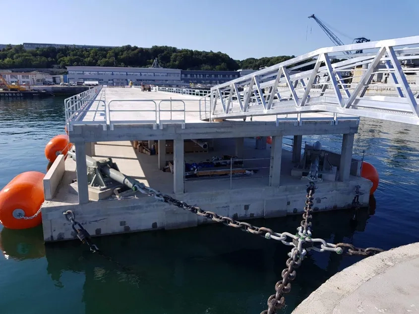 Les chaînes d’amarrage du ponton (ici les deux chaînes côté musoir) sont fixées à celui-ci via des amortisseurs (en gris) dimensionnés pour reprendre les efforts d’accostage et d’amarrage des frégates. Pour supporter ces charges élevées, les amortisseurs reposent sur des socles en béton très fortement armé. Les chaînes d’amarrage du ponton (ici les deux chaînes côté musoir) sont fixées à celui-ci via des amortisseurs (en gris) dimensionnés pour reprendre les efforts d’accostage et d’amarrage des frégates. Pour supporter ces charges élevées, les amortisseurs reposent sur des socles en béton très fortement armé.