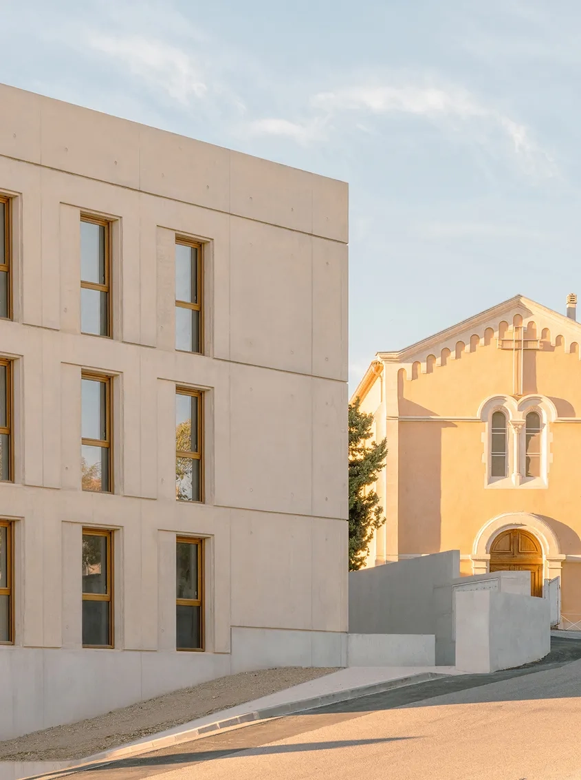 Le bâtiment affiche un béton beige teinté dans la masse qui reprend la couleur des appareillages principaux de l’église voisine.