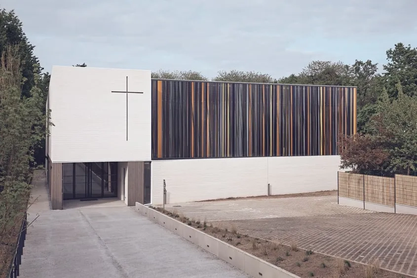 La façade de l’église décline une géométrie épurée dont les lignes horizontales et verticales dessinent une composition de quatre surfaces orthogonales. Deux rectangles revêtus de briques de béton blanches, l’un ancré au sol, l’autre soulevé, se touchent à leur seul angle droit commun. Sous le second, une ouverture en creux invite à entrer.