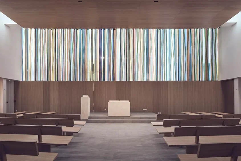 Placé au-dessus de l’autel, le grand vitrail de 120 m2 composé de verres profilés multicolores irradie d’une lumière frontale le sanctuaire.