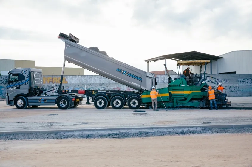 Un camion-benne, bâché durant le transport du BCR, déverse le matériau dans le finisseur. Celui-ci répand, nivelle et compacte le BCR.